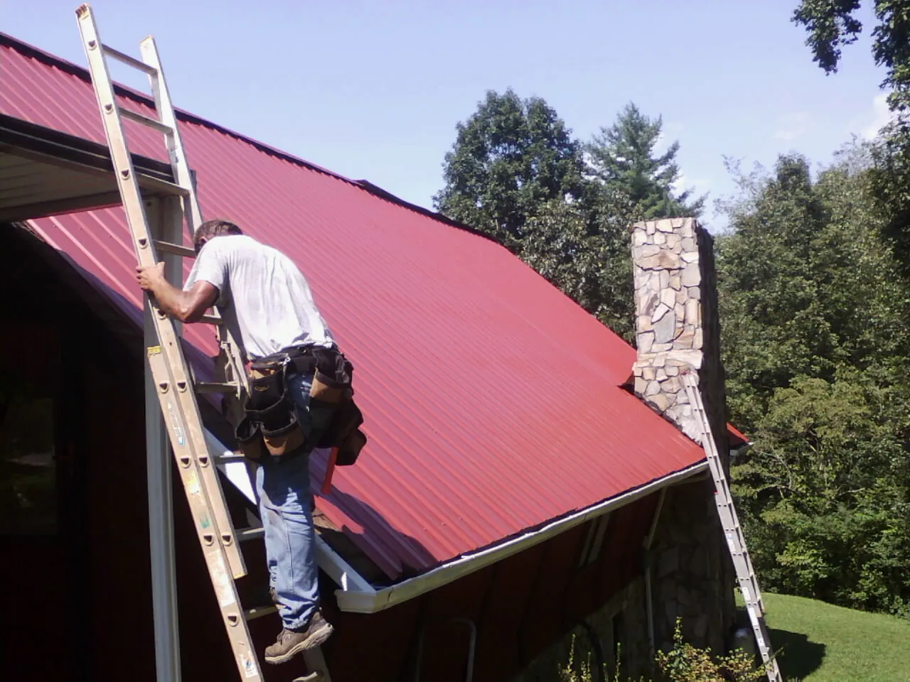Roofer on a ladder beside a red metal roof and stone chimney — Soffit & Fascia Repair by Roofing Brothers in Grandy